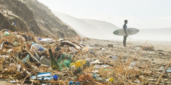 A man with surfboard stands on a beach littered with rubbish